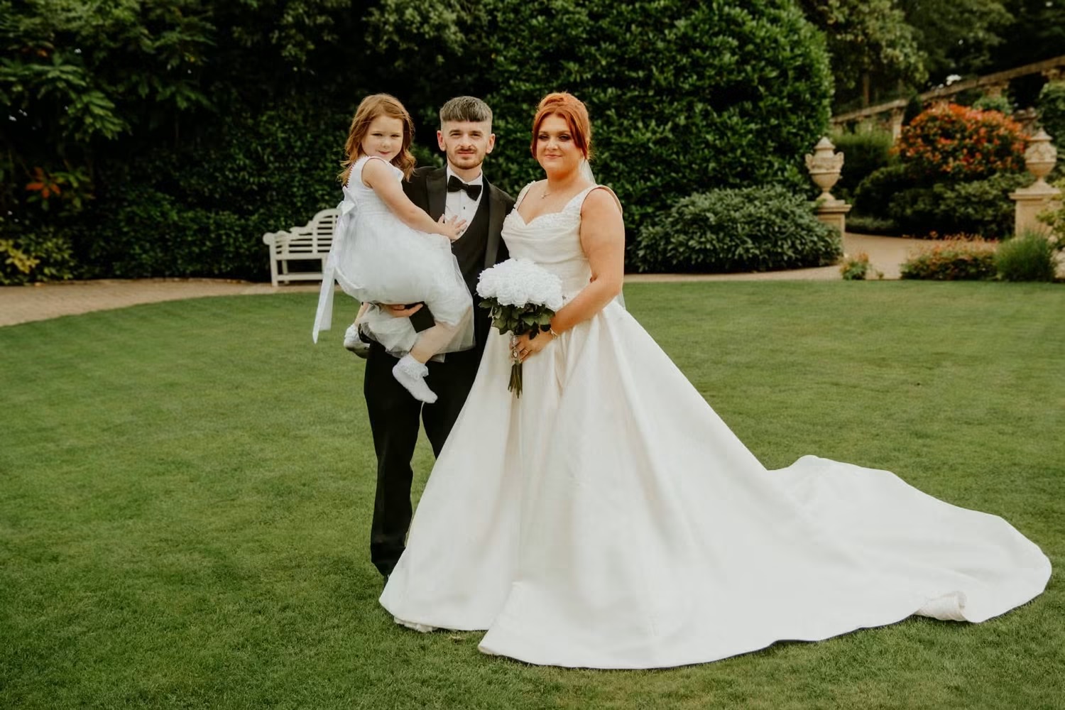Bride and groom pose in wedding attire, smiling warmly, with the bride holding a bouquet of white and green flowers.