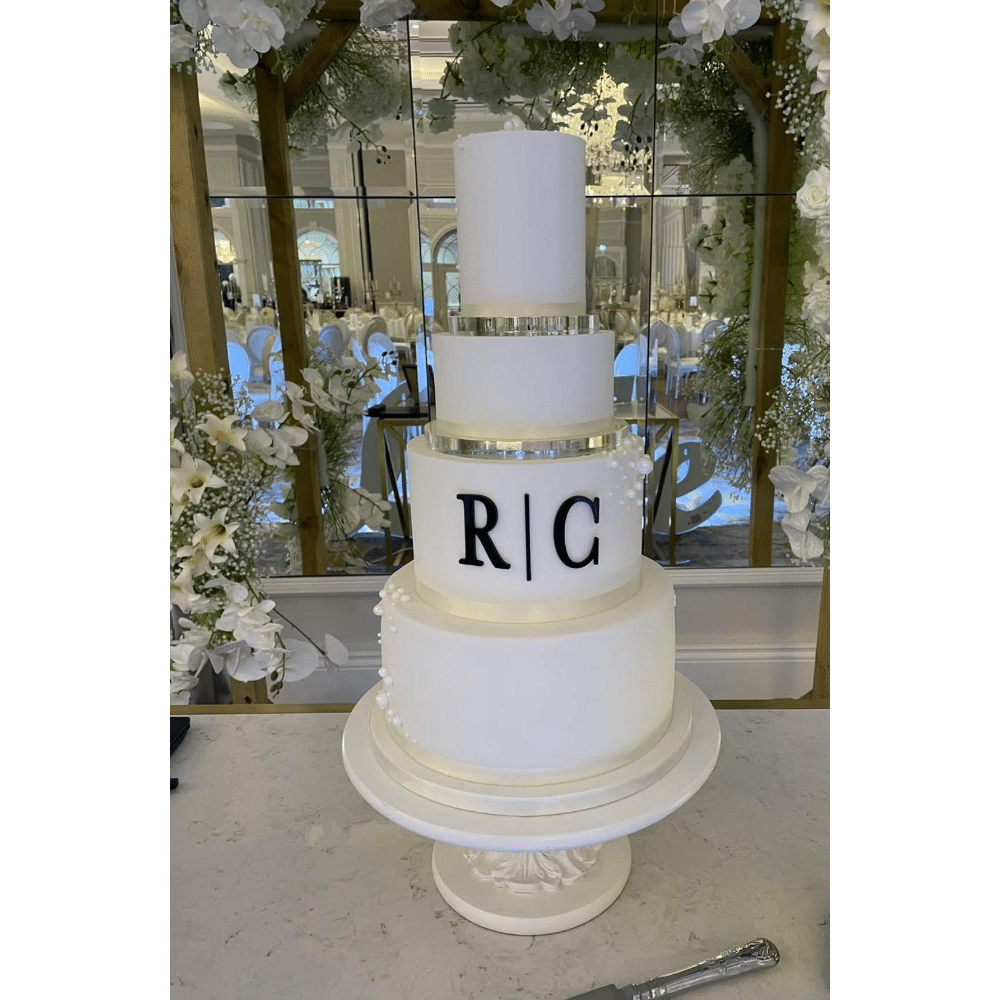 Elegant four-tier white wedding cake with initials "R | C," adorned with pearls, set in a decorated venue.