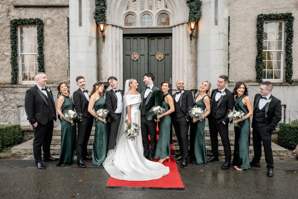 Wedding party posing on red carpet at Bellingham Castle, with bridesmaids in green dresses and groomsmen in black suits.