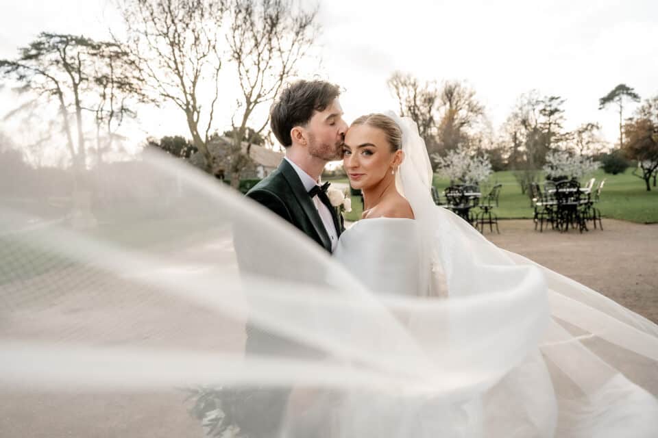 Bride and groom embrace outdoors with veil flowing at Bellingham Castle.