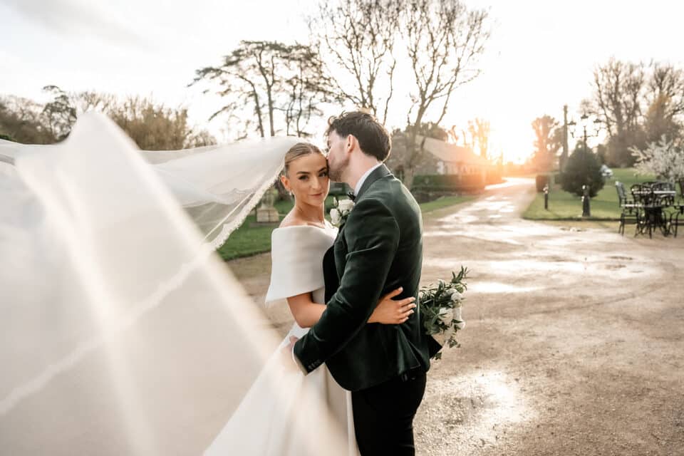 Bride and groom embrace at sunset in front of Bellingham Castle, with bride's veil flowing and lush greenery around them.