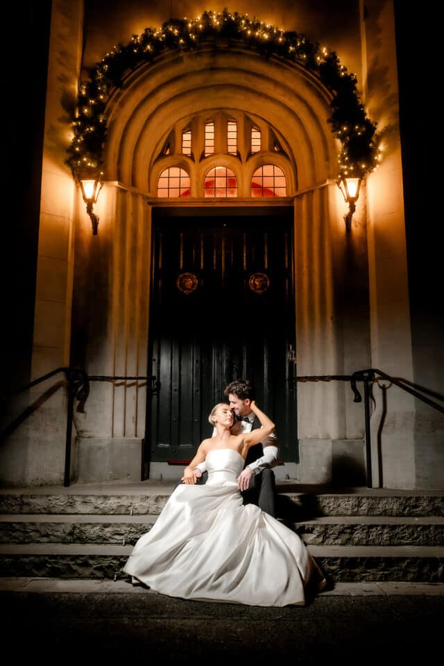 Bride and groom sitting on steps at night, Bellingham Castle entrance decorated with lights.