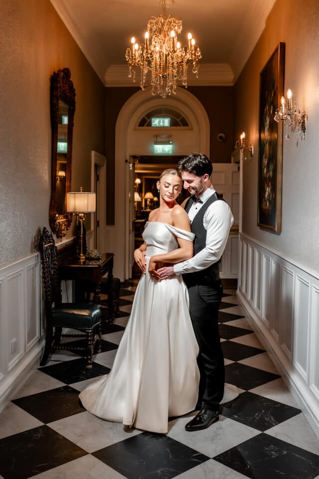 Bride and groom posing elegantly in a luxurious hallway with chandeliers and classic decor.