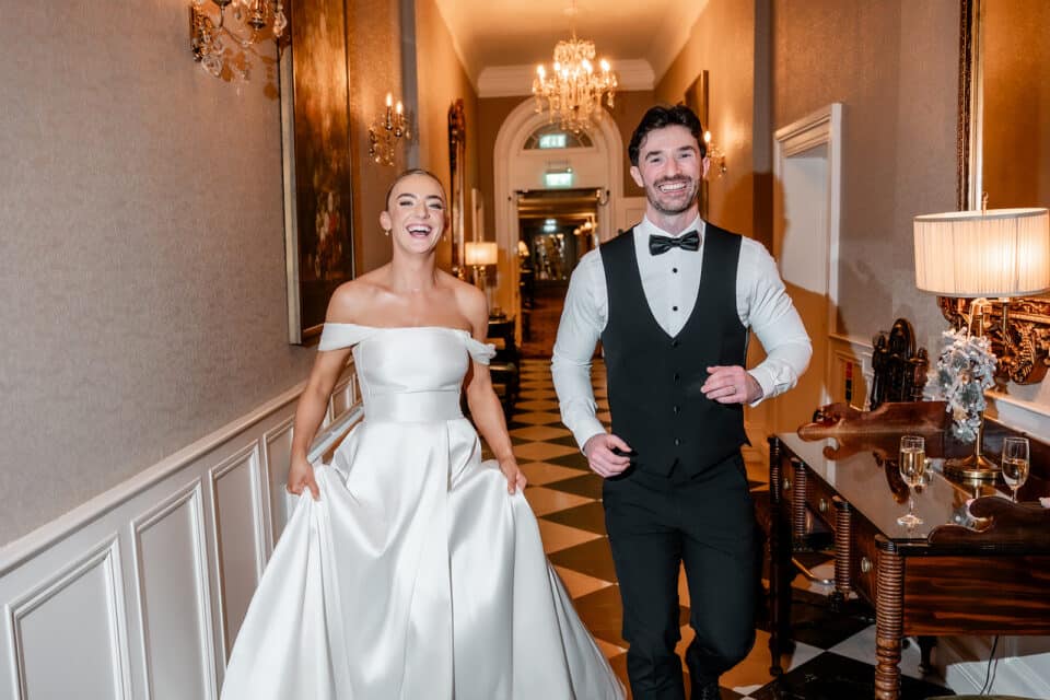 Bride and groom joyfully walking in a hallway at Bellingham Castle, dressed in wedding attire.
