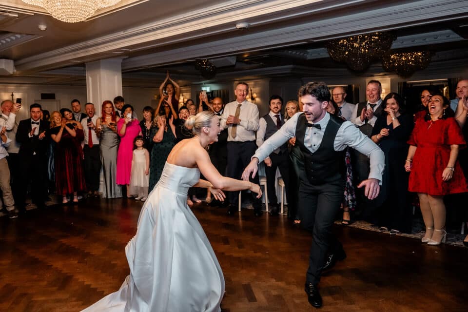 Bride and groom dancing joyfully at wedding reception with guests cheering in elegant venue.