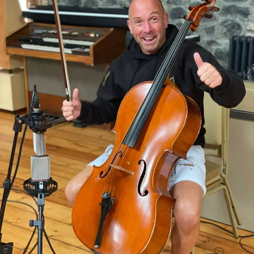 Man smiling while playing a cello, giving a thumbs-up, in a music studio setting.