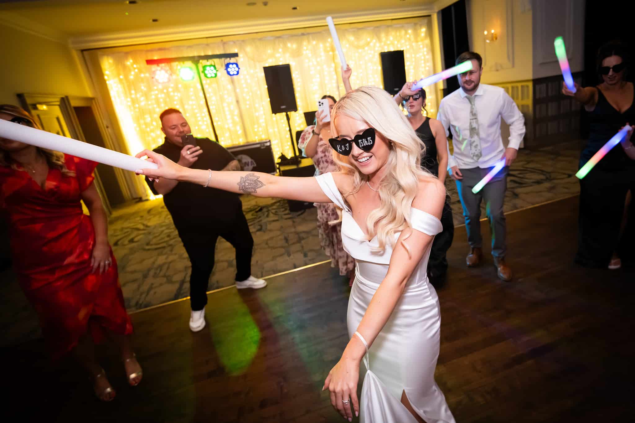A bride dances joyfully with friends at a wedding party, holding glow sticks under festive lighting.