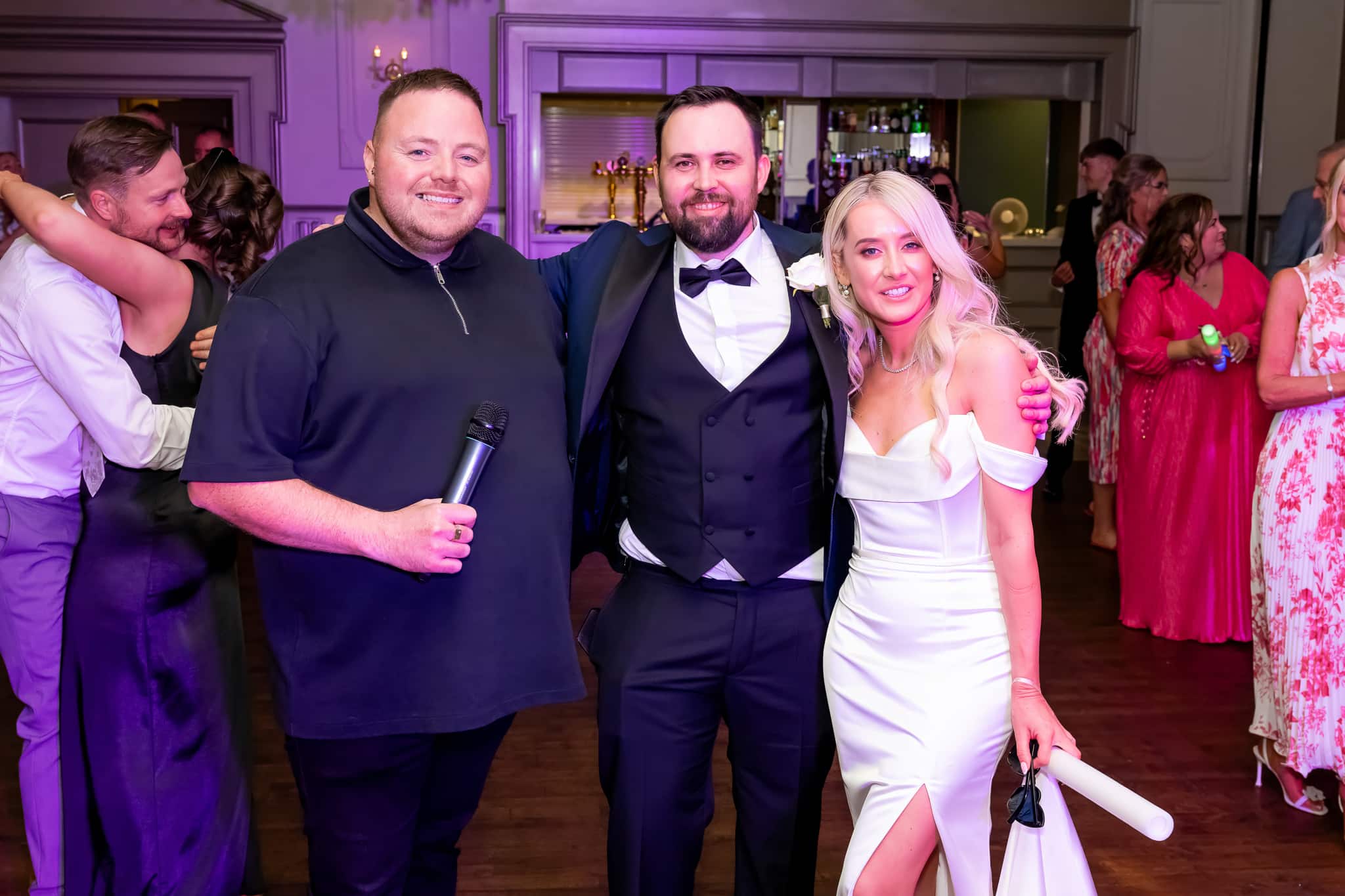 Group photo at a wedding reception with people posing and smiling, one holding a microphone.