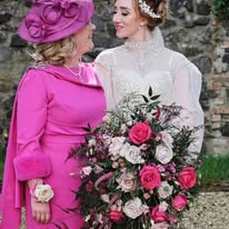 Bride in white gown with large bouquet stands beside woman in pink dress and hat, both smiling at each other.