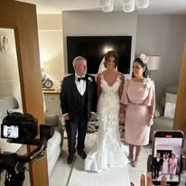 Bride in lace gown posing with parents in a living room, surrounded by cameras.