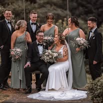 Bride in white gown with groom and bridal party, all smiling, in outdoor setting.