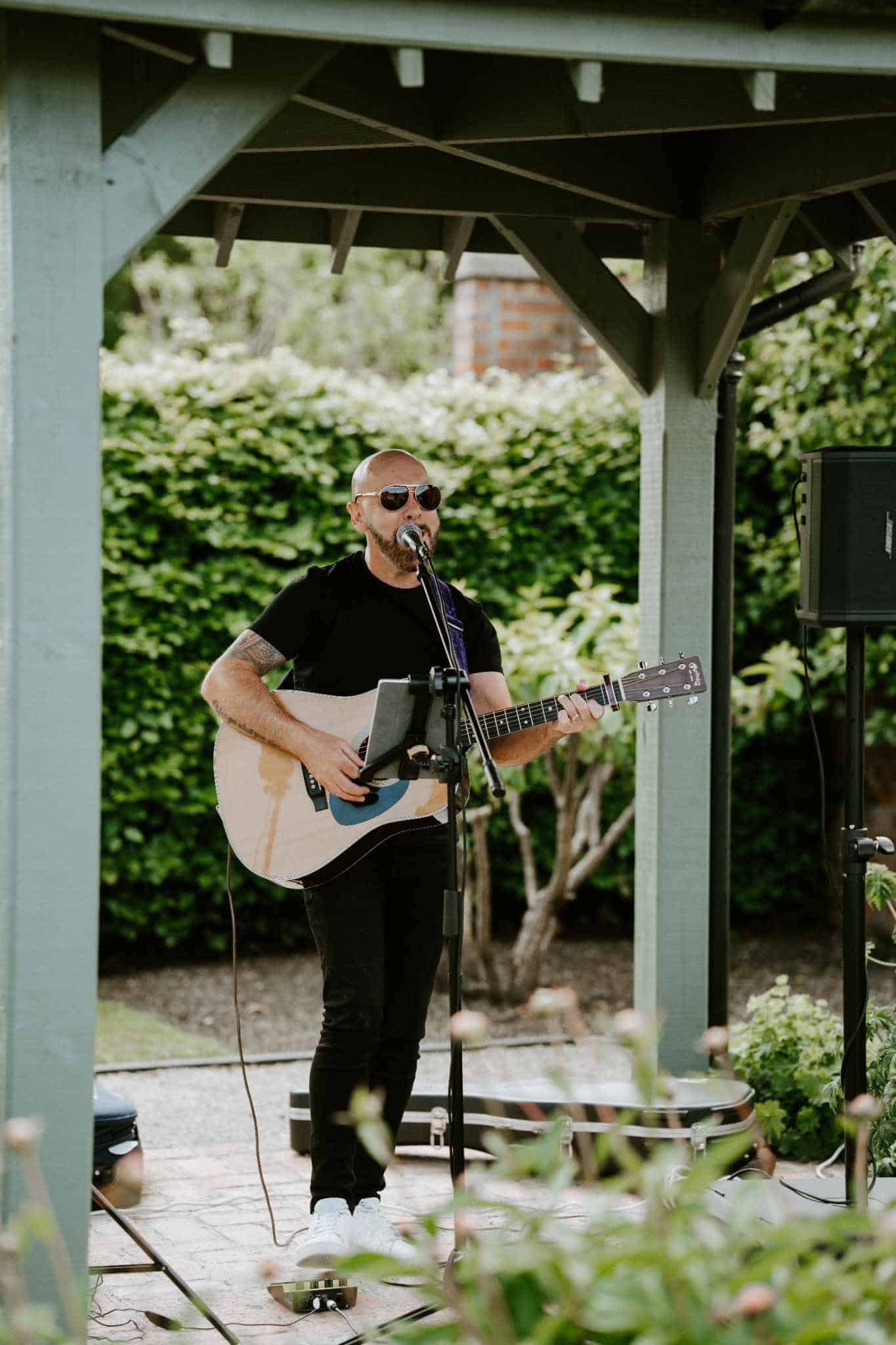 Musician playing guitar and singing outdoors under a gazebo.